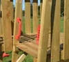 A close-up view of a wooden playground structure with red handles, set against a blurred outdoor background of grass and trees. Podgląd zdjęcia produktu
