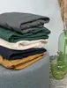 A stack of folded sheets in various colors sits on a gray ottoman next to two green glass vases. Produktbild-Vorschau