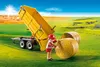 A toy farmer is pushing a hay bale next to a yellow trailer with the bed tilted up, in a field with hay bales in the distance. Voir les images du produit