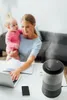 A woman is holding a baby while working on a laptop, with a humidifier in the foreground. Podgląd zdjęcia produktu