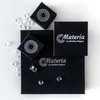 Schwarze Schmuckverpackungen mit Materia-Logo, dekorativ arrangiert mit klaren Acrylsteinen, zur Präsentation der Produktverpackung. Produktbild-Vorschau