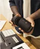 A person is holding two parts of a black Bluetooth speaker over a wooden surface with a keyboard and a laptop in the background. Náhled obrázku produktu