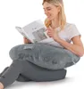 A woman is sitting on the floor, reading a book while using a U-shaped pillow for support. Produktbild-Vorschau