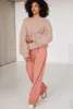 A woman models a beige sweater and pink pants in a studio setting. Produktbild-Vorschau