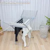A heated clothes drying rack with laundry on it, placed on a wooden floor in front of a white curtain. A laundry basket is visible to the right. Produktbild-Vorschau