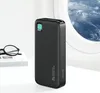 A black power bank is placed on a white surface, with a pair of glasses and a book to its left. In the background, an airplane window reveals a view of mountains and clouds. Náhled obrázku produktu