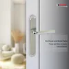 A close-up of a silver door handle with a brushed metal plate, set against a blurred background of a room interior and a reflective door surface. Produktbild-Vorschau