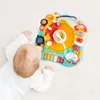 A baby lying on a white carpet, looking at a colorful interactive music toy. Produktbild-Vorschau