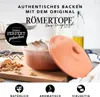 A terracotta bread baking pot with its lid slightly ajar, placed on a wooden board with some ingredients. The background shows a kitchen setting with a bottle of oil and a scale. Produktbild-Vorschau