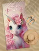A beach towel with a unicorn design is laid out on the sand, next to a straw hat and flip-flops. Anteprima dell’immagine prodotto