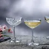Two crystal champagne glasses filled with champagne, placed on a wooden table with flowers in the background. Náhled obrázku produktu