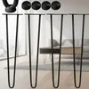 Four black hairpin legs are displayed in the foreground, with a blurred background showing a living room setting. Náhled obrázku produktu