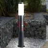 A black outdoor lamp with a motion sensor is shown in a garden setting, surrounded by gabion walls filled with stones and gravel. Produktbild-Vorschau