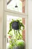 A hanging plant pot with a trailing plant is shown in front of a window, with a balcony and buildings visible outside. Náhľad obrázku produktu