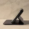 A black leather iPhone case with a red stitch detail is shown in a side profile, propped up in its stand function. Produktbild-Vorschau