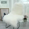 A white faux sheepskin rug draped over a chair, with a framed picture and a potted plant in the background. Produktbild-Vorschau