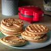 A red waffle maker is in the background with stacks of waffles in the foreground. Produktbild-Vorschau
