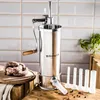 A stainless steel sausage stuffer with a wooden handle, along with raw sausages and stuffing tubes, set against a tiled background and wooden surfaces. Produktbild-Vorschau