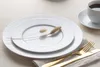 A white dinner plate set on a light-colored tablecloth, with a cup of coffee and gold cutlery nearby. Produktbild-Vorschau