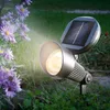 A solar-powered spotlight illuminates purple flowers in a garden at dusk. Produktbild-Vorschau