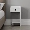 A white bedside table with a black metal leg is placed next to a bed in a room with gray walls and white baseboards. Produktbild-Vorschau