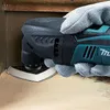A gloved hand is using a Makita oscillating multi-tool to sand a corner of a wooden surface. Produktbild-Vorschau