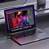 A laptop is open and displaying an image editing software with a photo of a person in a red jacket and yellow hat. The laptop is connected to a cable. Produktbild-Vorschau