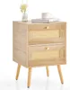 A light wood nightstand with two rattan-front drawers and gold knobs, shown against a white background. A small vase with a white flower and a book with a cup on top are placed on the surface. Produktbild-Vorschau