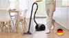 A person is vacuuming a wooden floor in a dining room area with chairs and a table visible. A German flag emblem is in the lower right corner. Produktbild-Vorschau