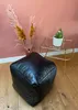 A black leather pouf sits on a wooden floor next to a small gold side table with a potted cactus on it. Dried pampas grass in a vase is behind the pouf. Produktbild-Vorschau