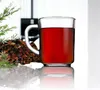 A clear glass mug filled with a red beverage, placed on a reflective surface next to a pile of dried herbs or spices and a sprig of greenery. Produktbild-Vorschau