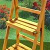 A close-up view of a wooden plant stand with multiple shelves, set against a blurred background of flowers and greenery. Produktbild-Vorschau