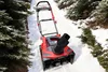 A red and black snow blower is on a snow-covered path between trees. Produktbild-Vorschau