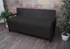 A brown poly-rattan storage bench with a black cushion sits on a wooden floor in front of a wood-paneled wall. A potted plant is visible to the right of the bench. Náhľad obrázku produktu
