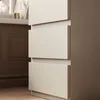 A close-up view of a beige dresser with three drawers, set against a brown wall and wooden floor. Produktbild-Vorschau