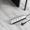 A power strip with four outlets and a plug lies on a light gray wooden floor, next to the leg of a dark piece of furniture. Náhľad obrázku produktu