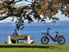 A person sits on a bench by a lake, listening to music, with an e-bike parked nearby. Produktbild-Vorschau