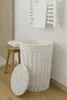 A white wicker laundry basket with a lid and a beige liner, placed on a wooden floor next to a small wooden stool with a folded white towel on it. A white towel rack with a towel is mounted on the wall behind the basket. Produktbild-Vorschau