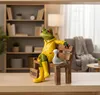 A decorative frog figurine sits on a wooden bench with a seagull, in a room with a bookshelf and armchair. Produktbild-Vorschau