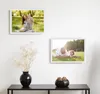Two framed pictures are displayed on a white wall above a black console table. The console table holds a black vase with dried flowers and a black bowl with decorative balls. The pictures show families in outdoor settings. Produktbild-Vorschau