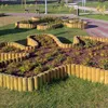 A park scene with curved wooden borders filled with plants and mulch, a paved path, and benches in the background. Produktbild-Vorschau