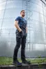 A man wearing overalls and a t-shirt stands in front of a large industrial silo. Náhled obrázku produktu