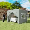 A man stands next to a gray pop-up gazebo set up in a grassy backyard with a brick wall and trees in the background. Inside the gazebo, a table is set with a white tablecloth and some decorative items. Podgląd zdjęcia produktu