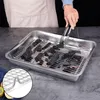 A person is using tongs to place charcoal pieces onto a metal cooling rack inside a metal baking tray. Produktbild-Vorschau