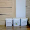 Three white trash cans are placed next to a wooden door and a white panel. Voir les images du produit