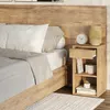 A bedside table next to a bed, with a lamp, clock, and books on it. Náhled obrázku produktu