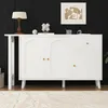 A white cabinet with arched doors and a drawer is displayed against a black wall, with a framed artwork above it and a lamp on its surface. Produktbild-Vorschau