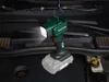 A cordless LED work light is hanging from a hook inside the open hood of a car. Produktbild-Vorschau