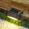 A solar-powered step light is mounted on a wooden deck, illuminating the surrounding grass and white gravel in the rain. Produktbild-Vorschau