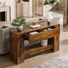 A wooden console table with two drawers is placed behind a sofa in a living room setting. The table has a rustic wood finish and is decorated with a plant, books, and a decorative radio. Produktbild-Vorschau
